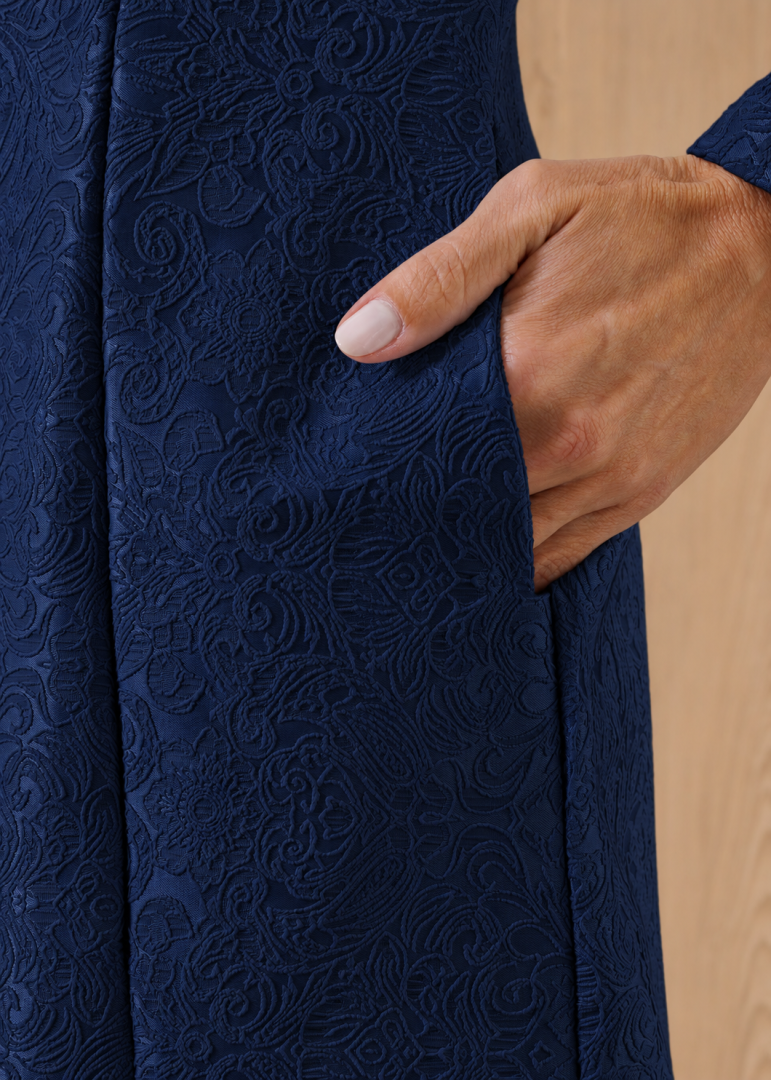 Close do bolso funcional do jaleco feminino azul-marinho, destacando o tecido jacquard com desenhos florais em relevo.